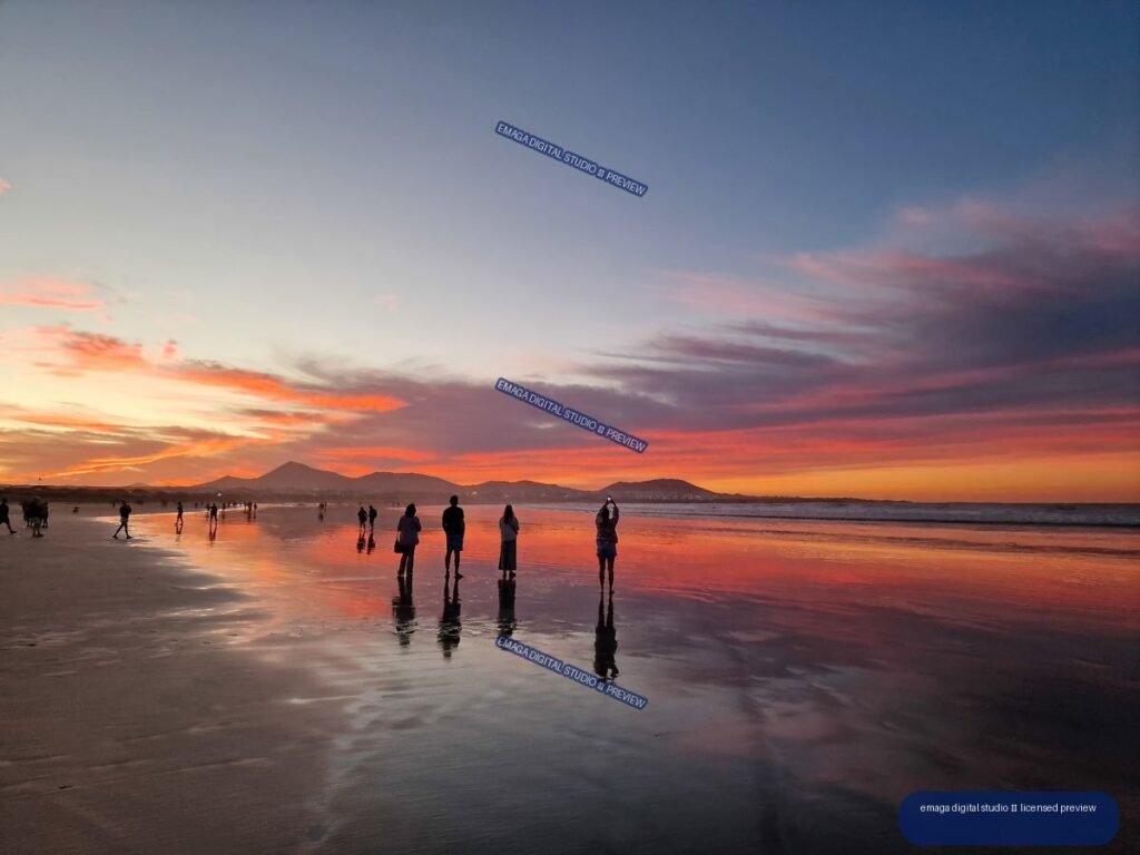People and their reflections on wet sand at Famara Beach beneath a vivid sunset, with mountains on the horizon.