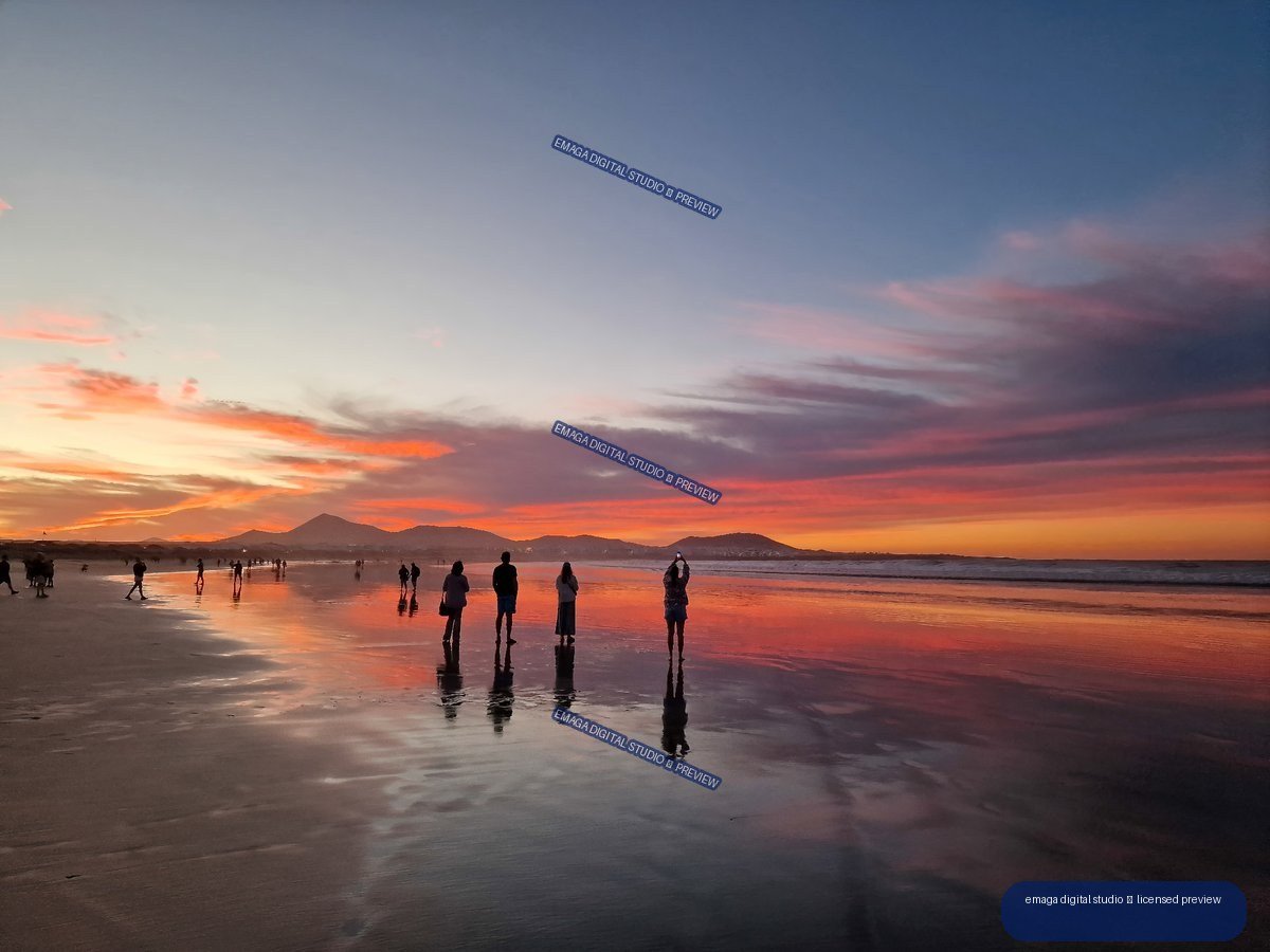 People and their reflections on wet sand at Famara Beach beneath a vivid sunset, with mountains on the horizon.