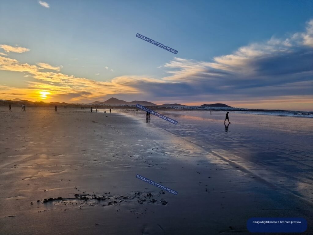 Sunset over Famara Beach with reflected light on wet sand, silhouetted people, distant volcanic hills, and a cloud-filled sky.