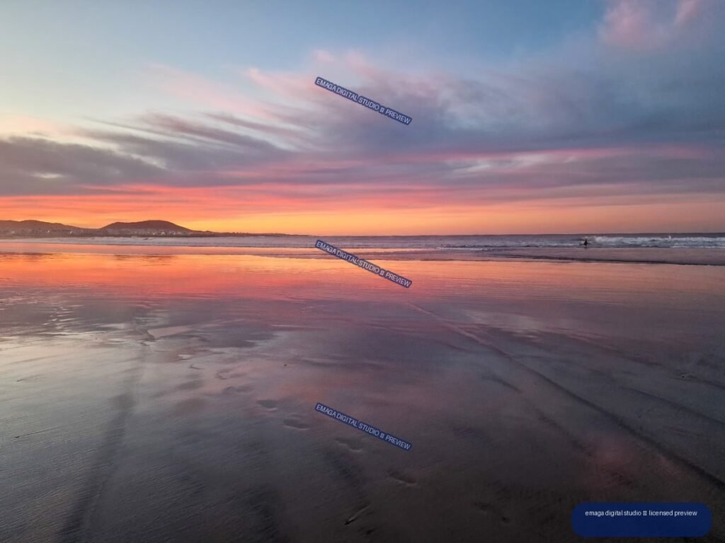 Sunset over a broad beach, with pink and purple clouds reflected in wet sand and shallow water; low hills sit on the distant horizon.