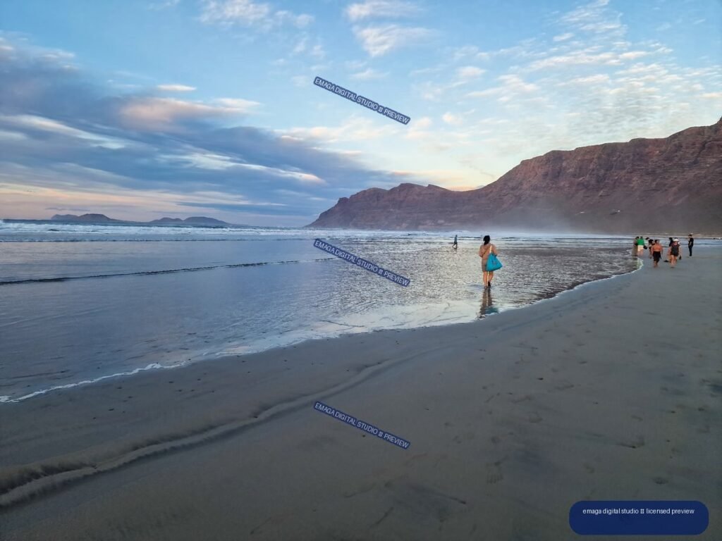 Wet reflective sand and gentle waves at Famara Beach, with people walking beneath dramatic cliffs and a cloud-filled evening sky.