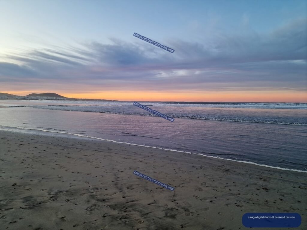 Pastel sunset over a broad sandy beach, with reflective shallows, gentle waves, a low headland, and distant buildings on the horizon.