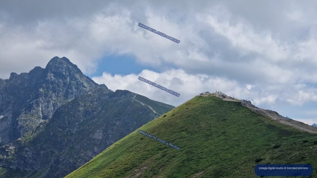Green alpine ridge with hikers on the crest, backed by a jagged gray mountain beneath a cloudy sky.