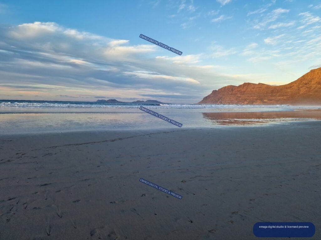 Wet reflective beach at sunset with waves, layered clouds, distant low islands, and sunlit reddish cliffs on the right.