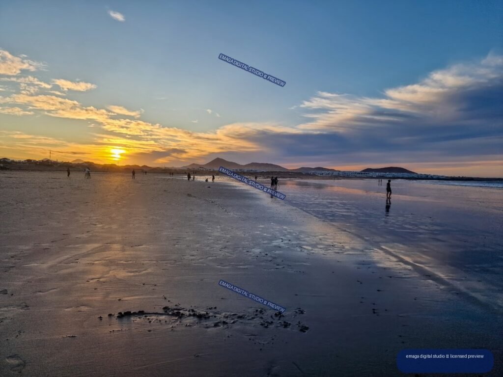 Sunset over Famara Beach with mountains, scattered beachgoers, and colorful reflections in wet sand and shallow water.