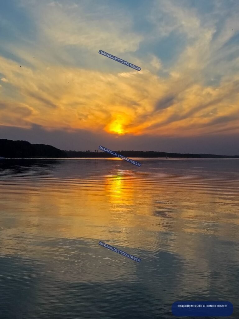 Golden sun setting behind a wooded shoreline, reflected in gently rippled lake water beneath streaked clouds.