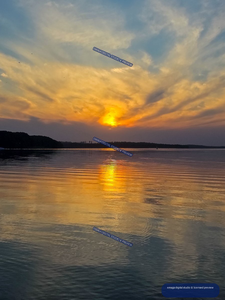 Golden sun setting behind a wooded shoreline, reflected in gently rippled lake water beneath streaked clouds.