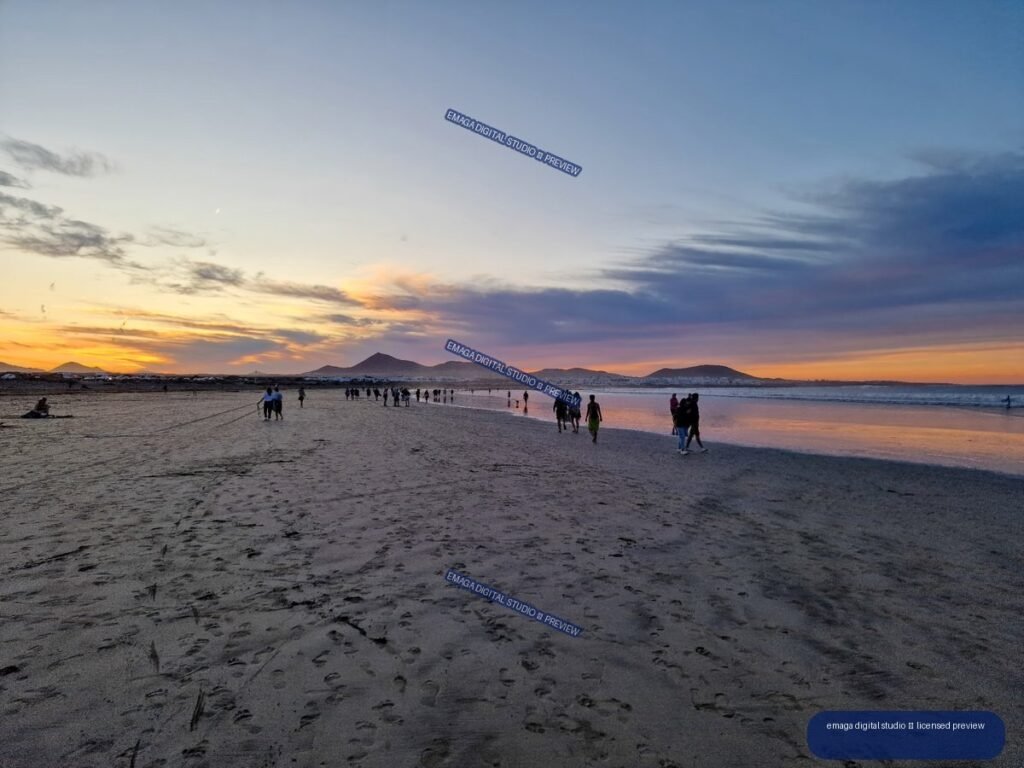 People walking across a broad sandy beach at sunset, with reflections, ocean water, and distant volcanic mountains beneath a colorful cloudy sky.