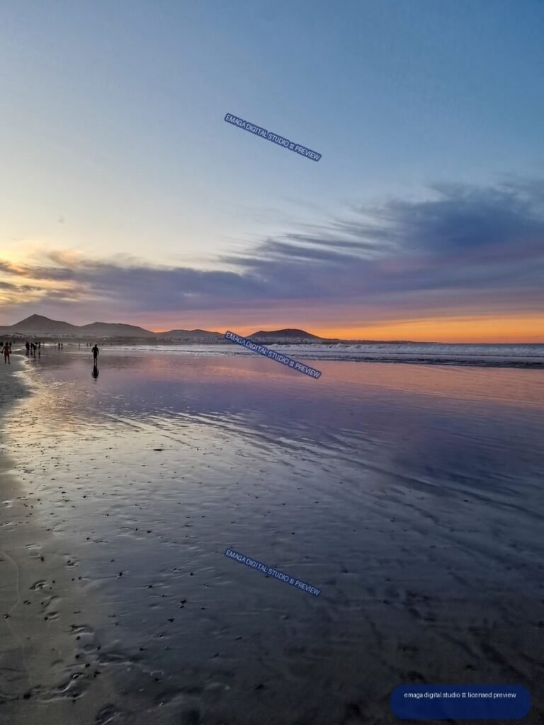Wet sand reflects a colorful sunset sky at Famara Beach, with waves, silhouetted people, and low hills on the horizon.