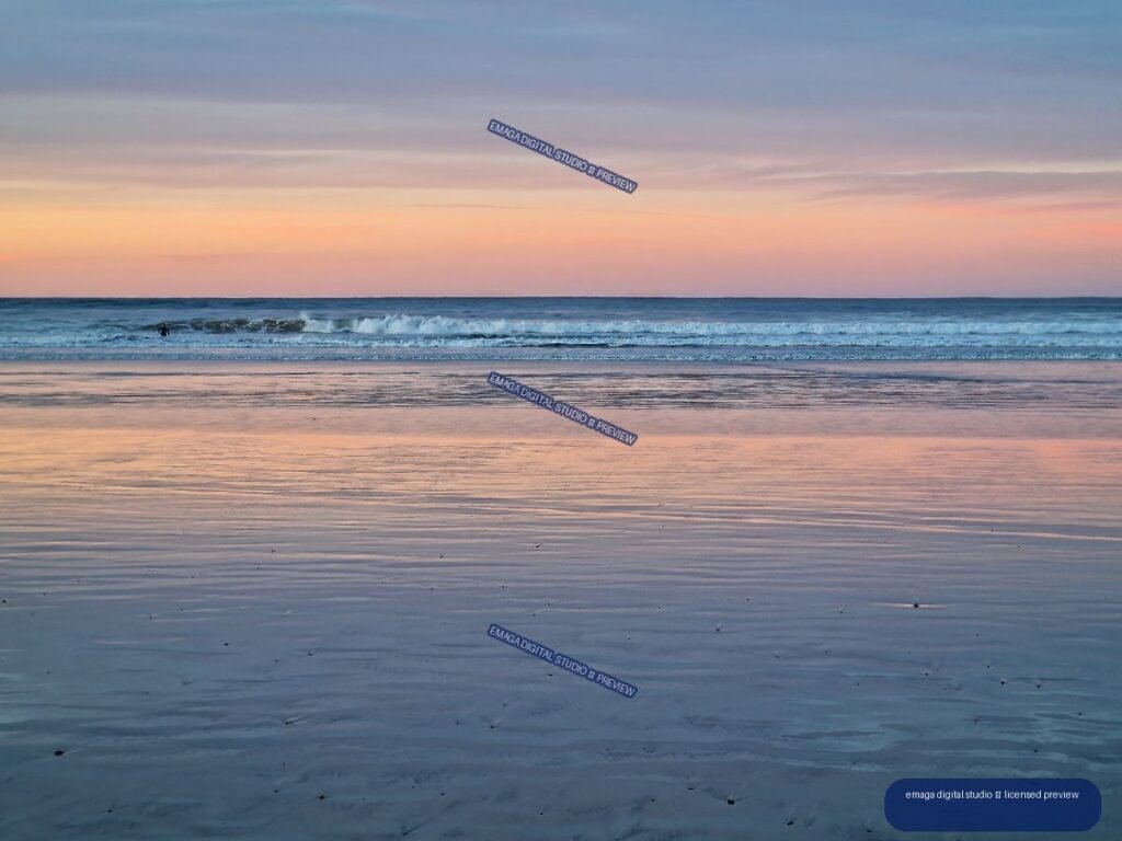 Pastel sunset reflected across wet sand at a beach, with gentle waves and a small figure near the surf.