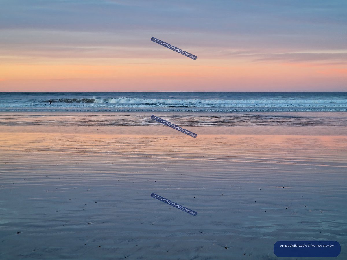 Pastel sunset reflected across wet sand at a beach, with gentle waves and a small figure near the surf.