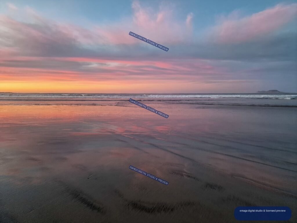 Sunset over a broad beach with pink and orange cloud reflections in wet sand and shallow water; low landform on the right horizon.