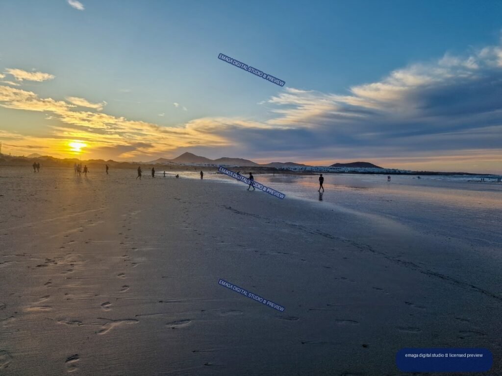 Sunset over a broad beach with reflective wet sand, distant volcanic hills, scattered people, and layered clouds.