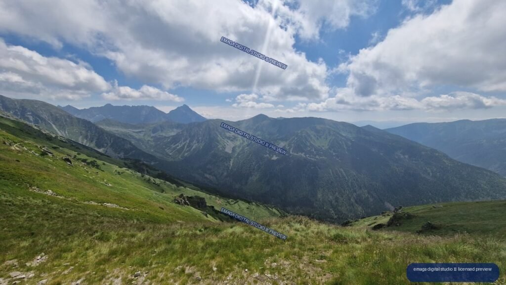Panoramic mountain landscape with a grassy rocky slope in the foreground, layered ridges and valleys beyond, and clouds in a blue sky.