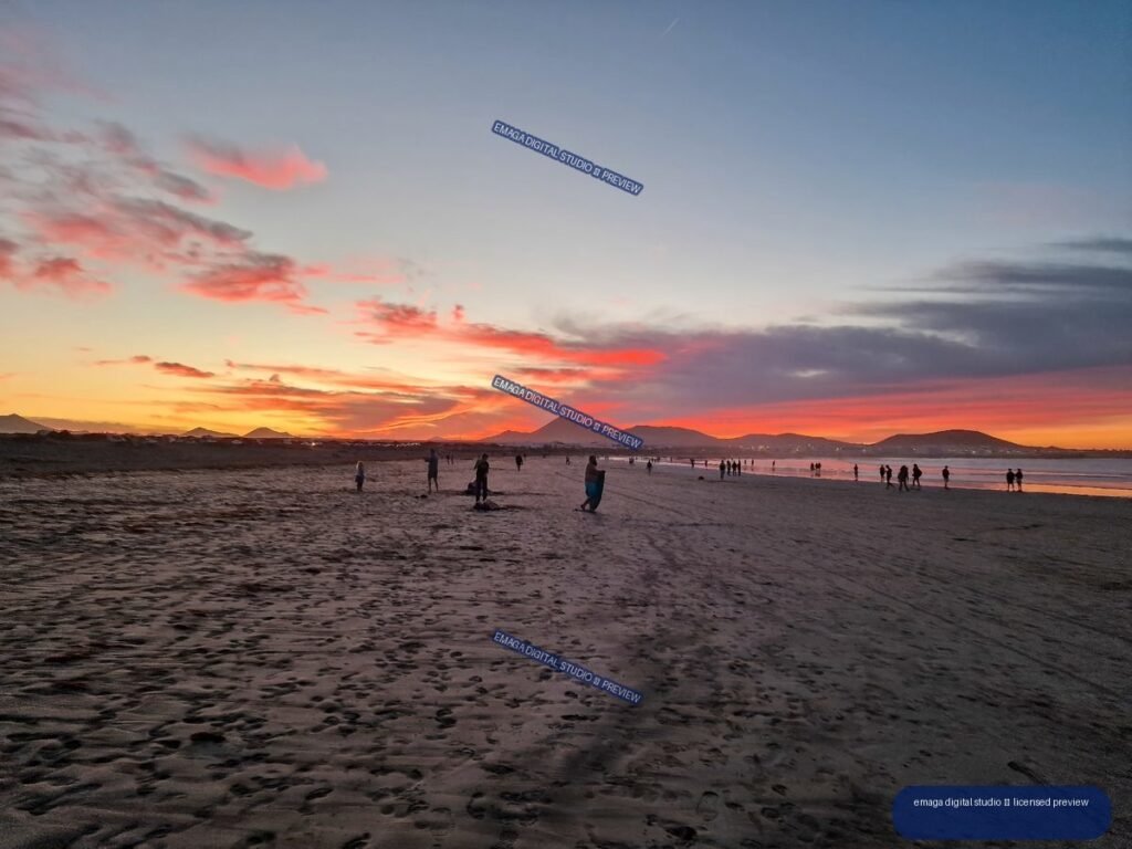 Sunset over Famara Beach with people on the shore, reflective wet sand, and distant volcanic mountains beneath colorful clouds.