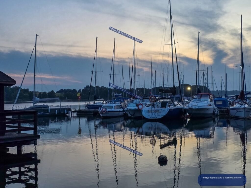 Sailboats and motorboats moored at a marina on a lake at dusk, reflected in calm water beneath a cloudy sky.