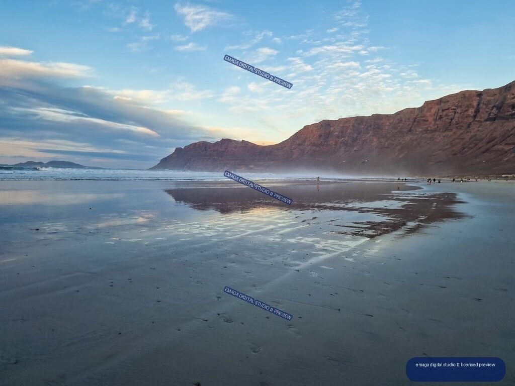 Wet beach sand reflects a blue and softly illuminated sky beneath rugged reddish cliffs, with waves and small figures along the shoreline.