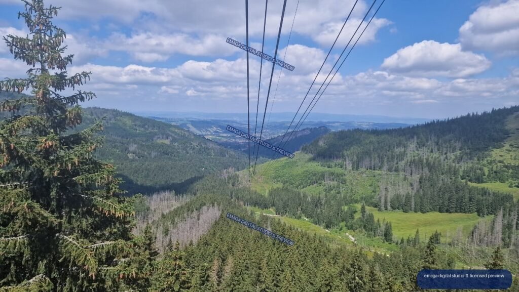 Cable-car lines lead over forested mountain slopes toward a distant valley and ridges under a blue, cloud-filled sky.