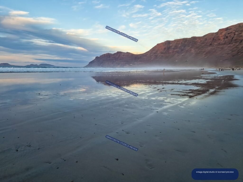 Reflective wet sand at Famara Beach beneath a cloudy evening sky, with waves, distant landforms, and a large reddish cliff.
