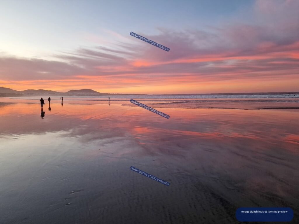Sunset over a beach with pink and lavender clouds reflected on wet sand, distant hills, waves, and small figures.
