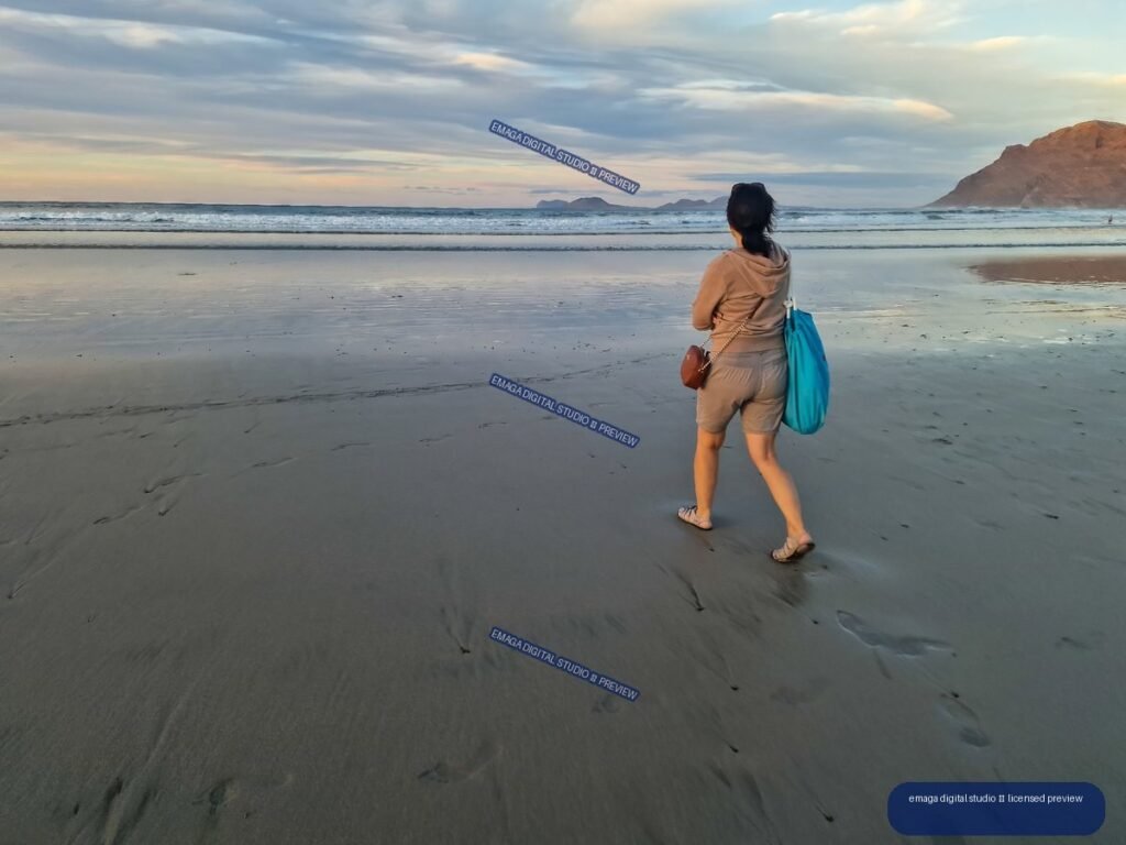 Woman with a turquoise bag walking across reflective wet sand toward the ocean at sunset, with waves, clouds, and volcanic cliffs.