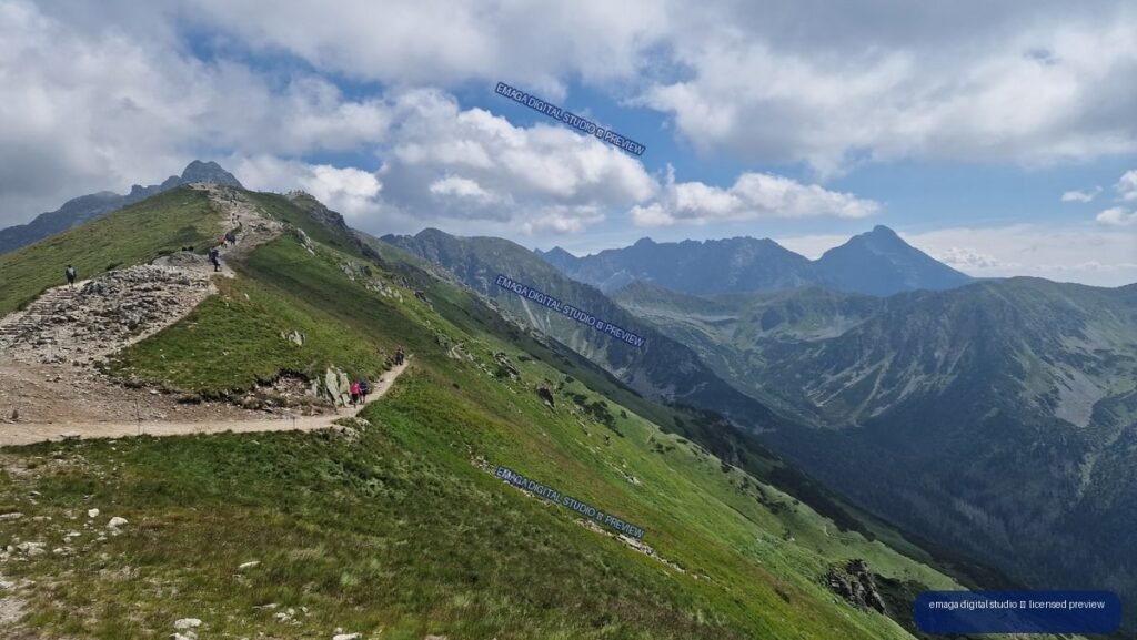 Hikers on a rocky trail crossing a green mountain ridge, with jagged peaks and valleys under a partly cloudy blue sky.
