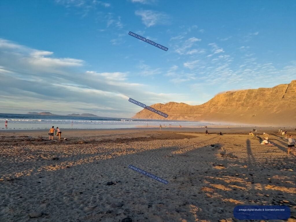 Wide view of Famara Beach with reflective wet sand, beachgoers, distant islands, and sunlit cliffs beneath a blue sky.