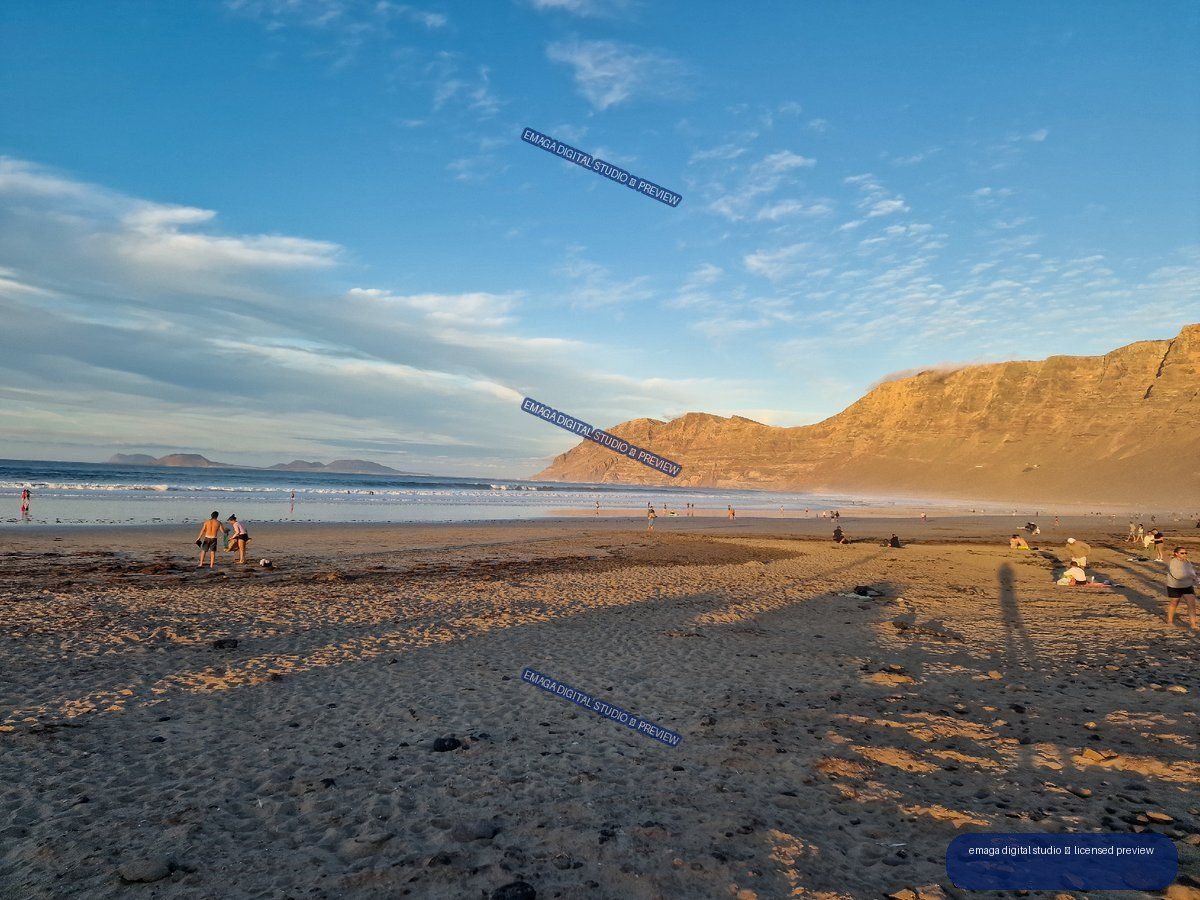 Wide view of Famara Beach with reflective wet sand, beachgoers, distant islands, and sunlit cliffs beneath a blue sky.