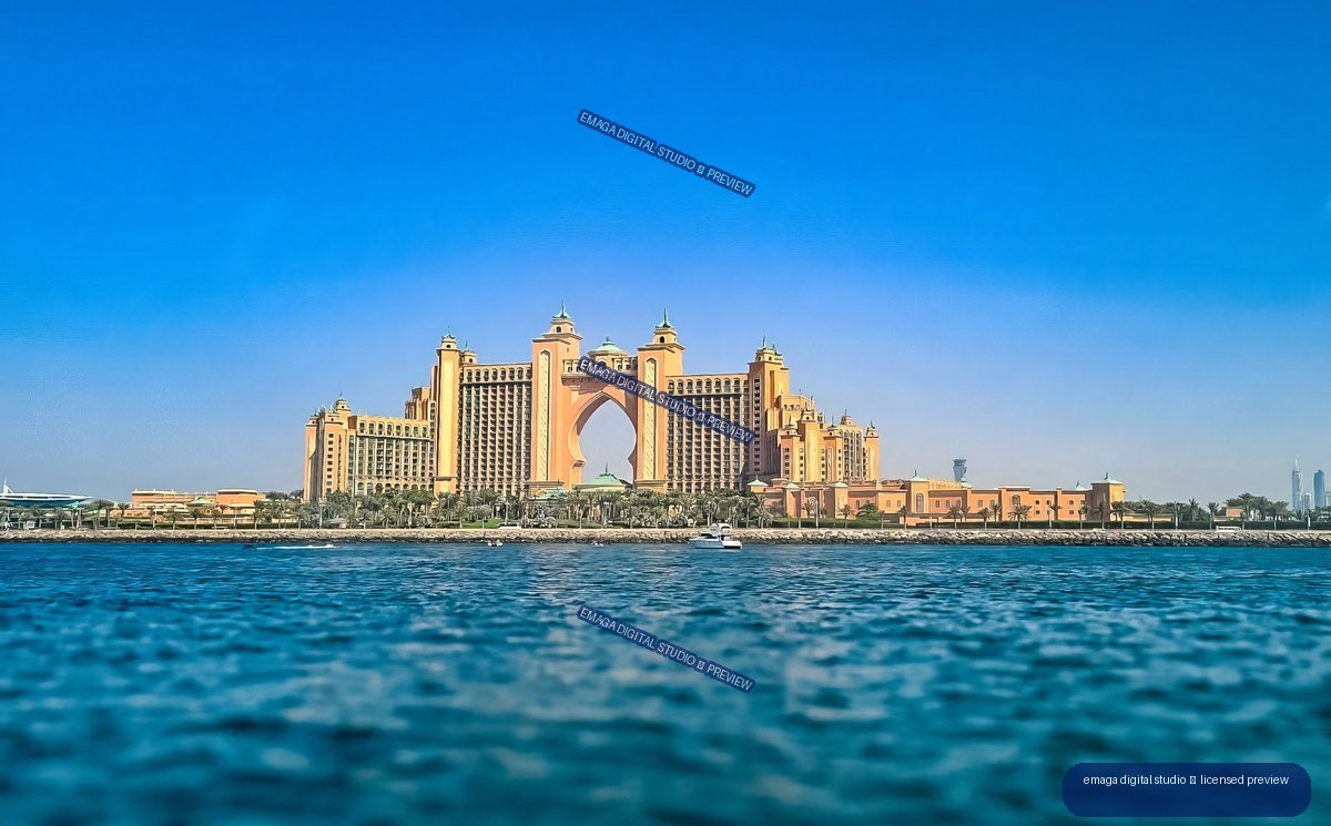 Atlantis The Palm resort viewed across rippling blue water under a clear sky, with Dubai skyscrapers faintly visible at right.
