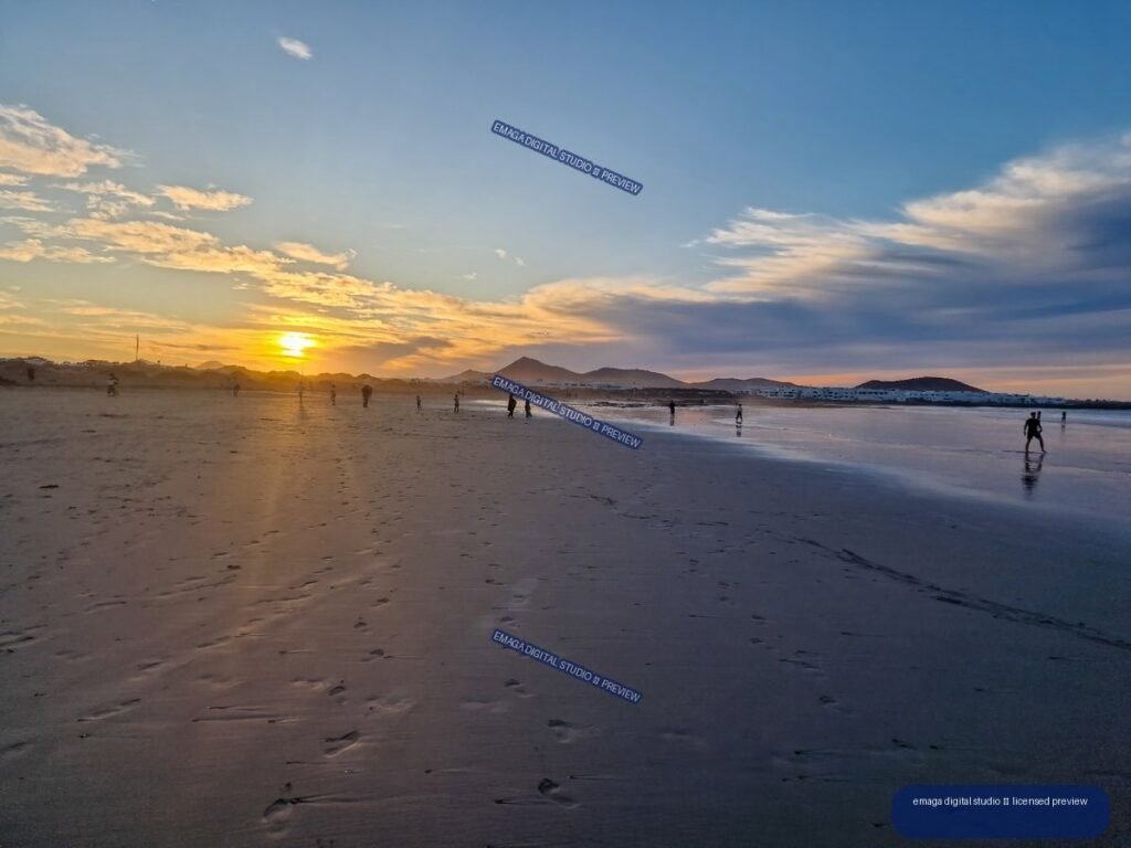 Sunset over a broad sandy beach, with reflected light on wet sand, silhouetted people, mountains, and layered clouds.