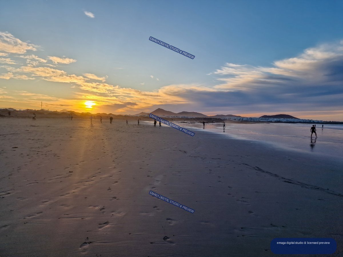 Sunset over a broad sandy beach, with reflected light on wet sand, silhouetted people, mountains, and layered clouds.