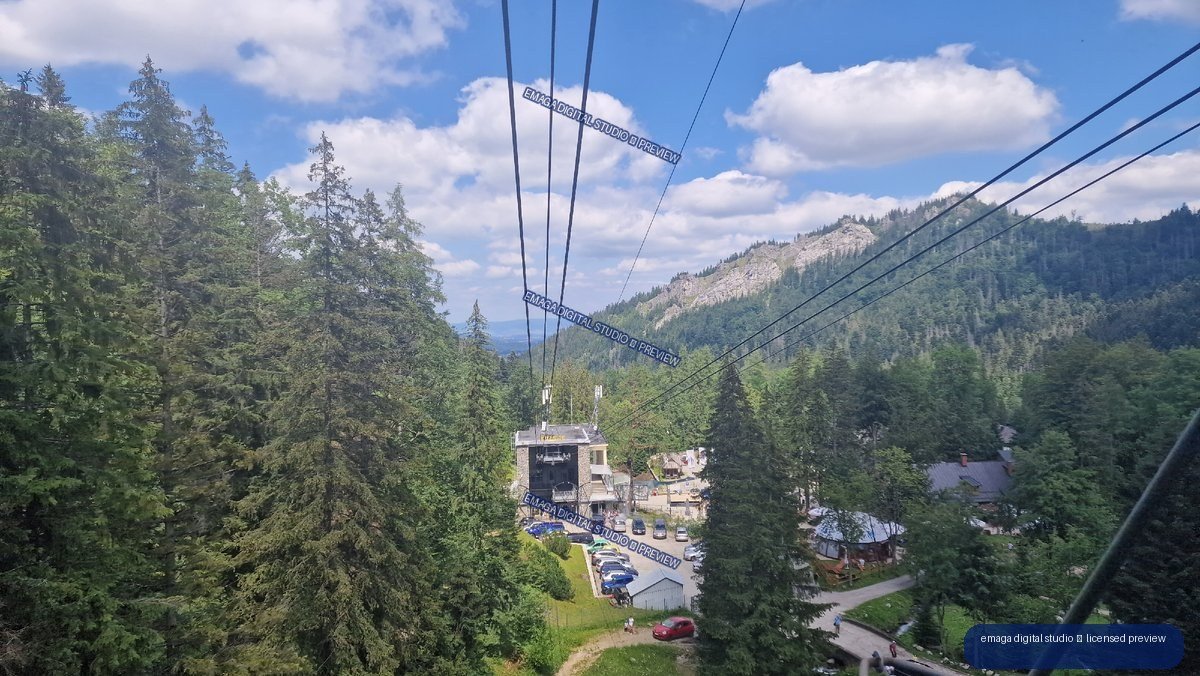 View from a cable car over a forested mountain station, with cables leading toward a rocky ridge under a blue, partly cloudy sky.