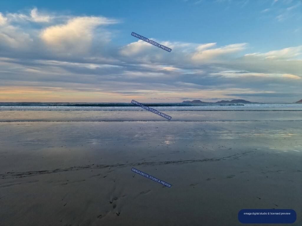 Wide view of wet reflective sand and gentle waves at Famara Beach beneath layered evening clouds, with distant mountains on the horizon.