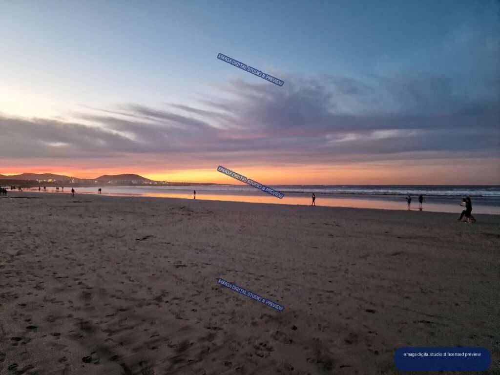 Wide sandy beach with people, ocean waves, distant hills, and orange-pink sunset light beneath layered clouds.
