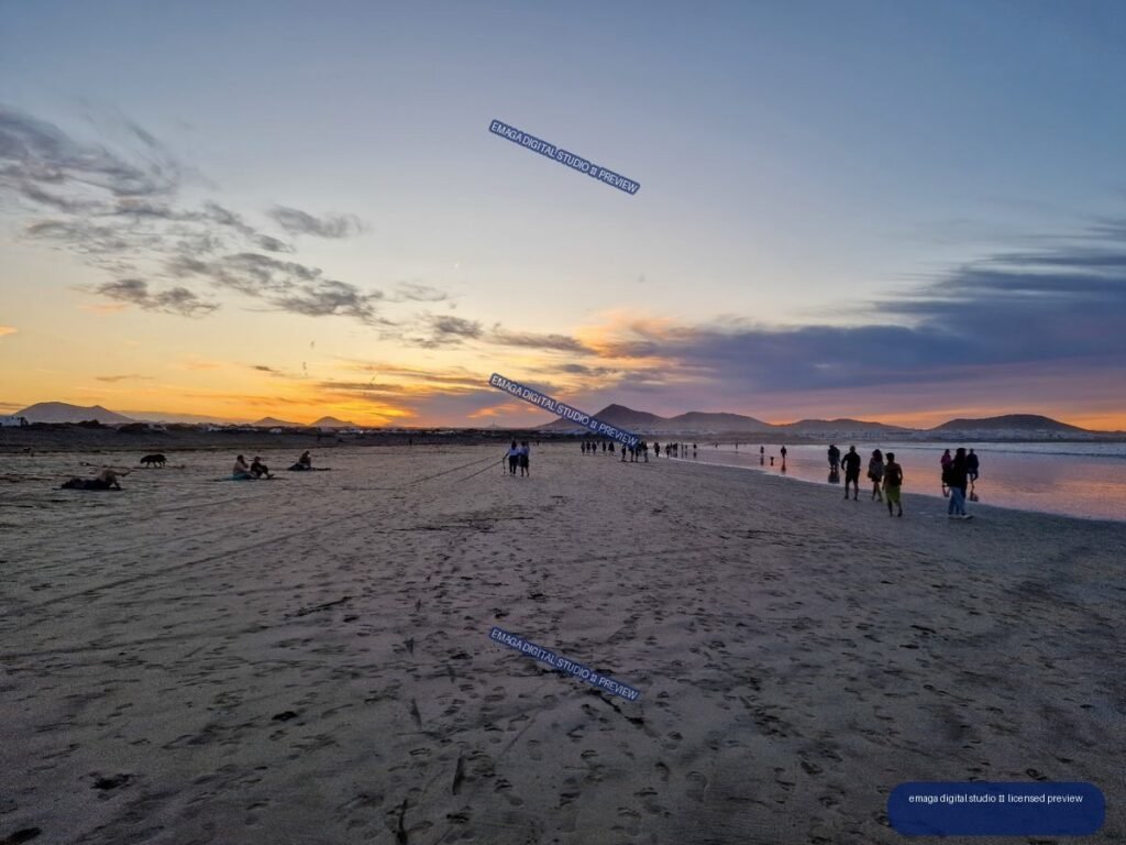 Sunset over Famara Beach with volcanic hills, reflective shoreline, clouds, and people walking across the sand.