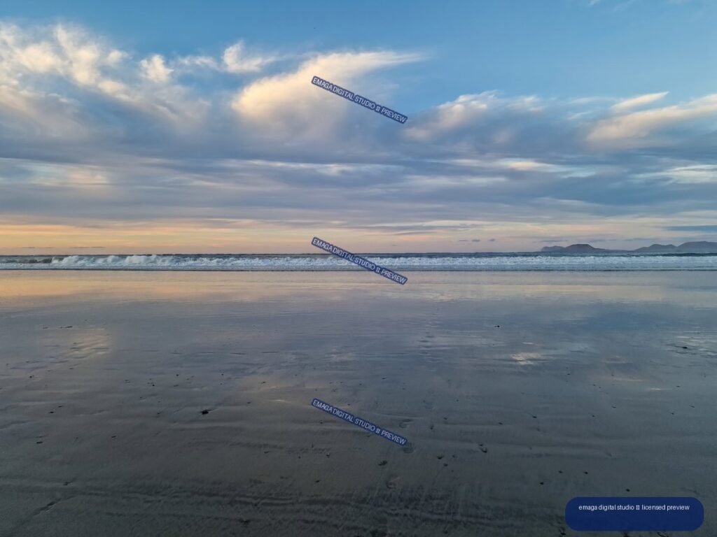 Reflective wet sand and breaking waves beneath layered clouds at sunset, with distant mountains on the horizon.