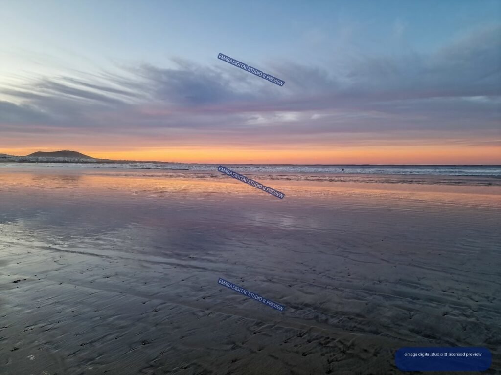 Sunset over a broad beach, with colorful clouds and the glowing sky reflected in wet sand and shallow water.