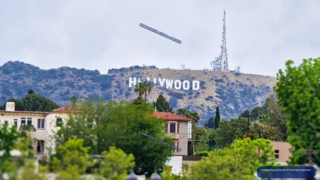 The Hollywood sign on a hillside above trees and Spanish-style houses, with communications towers on the ridge under an overcast sky.