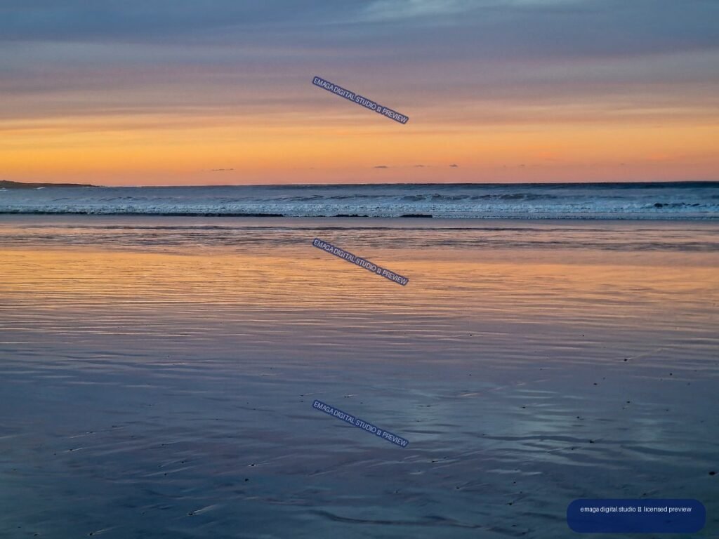 Wet beach sand reflecting an orange and blue sunset beneath a calm ocean horizon and gentle waves.