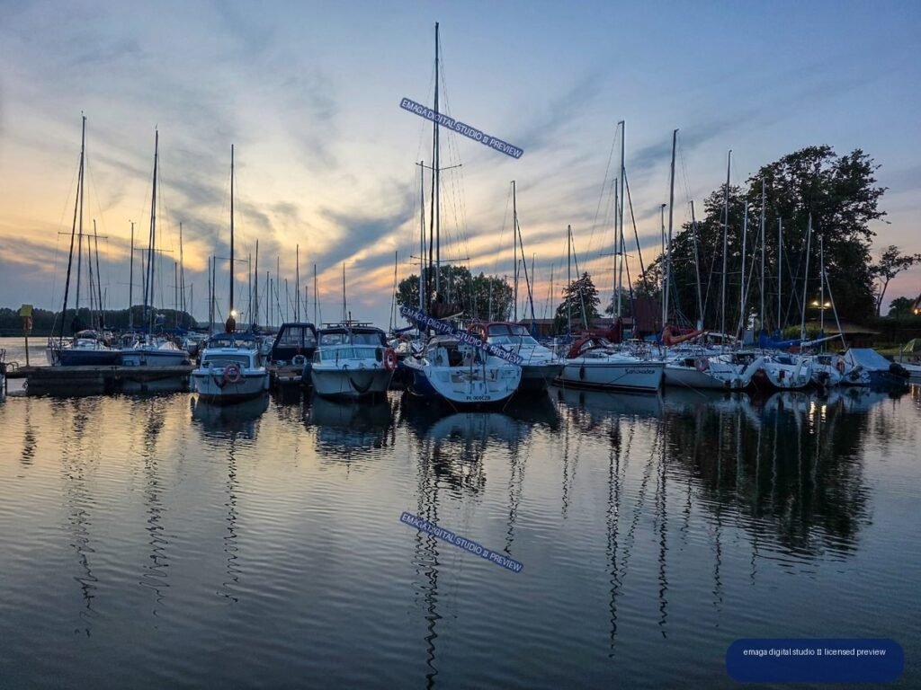 Sailboats and motorboats moored at a marina on calm reflective water beneath a cloudy sunset sky.