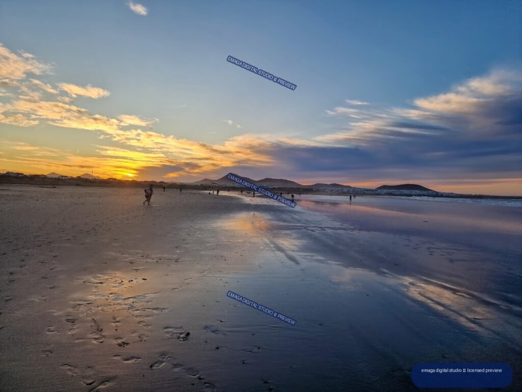 Sunset over Famara Beach with golden reflections in wet sand, distant volcanic hills, clouds, and small beachgoers.