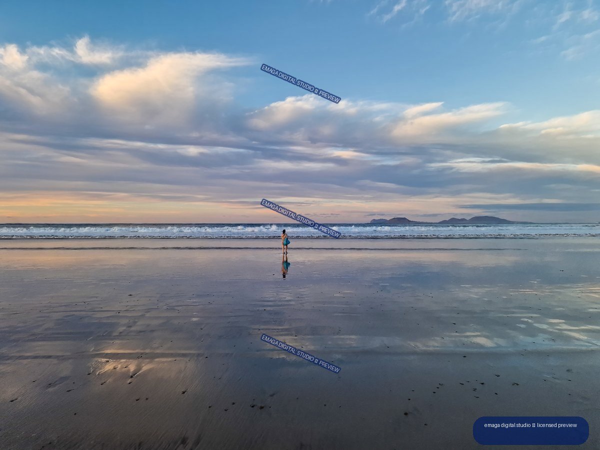A solitary person and their reflection stand on wet sand beneath layered clouds, with waves and distant mountains on the horizon.