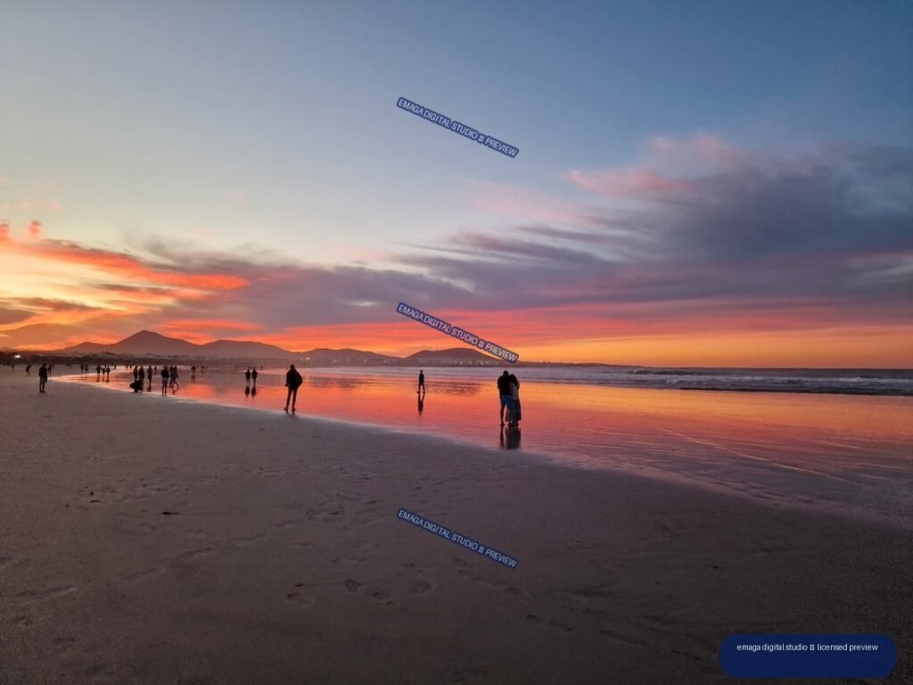 People on reflective wet sand at Famara Beach beneath a vivid sunset sky, with mountains on the horizon.