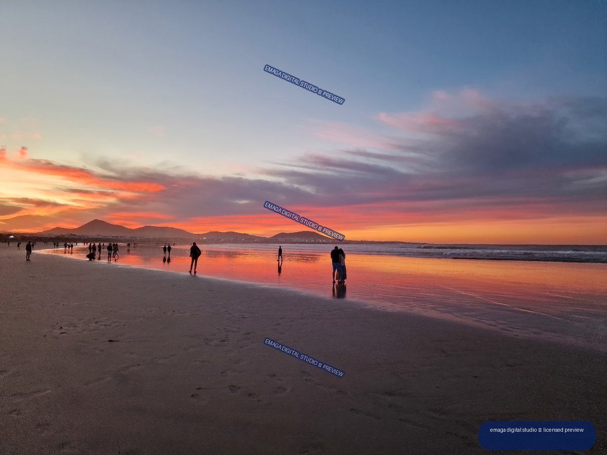 People on reflective wet sand at Famara Beach beneath a vivid sunset sky, with mountains on the horizon.