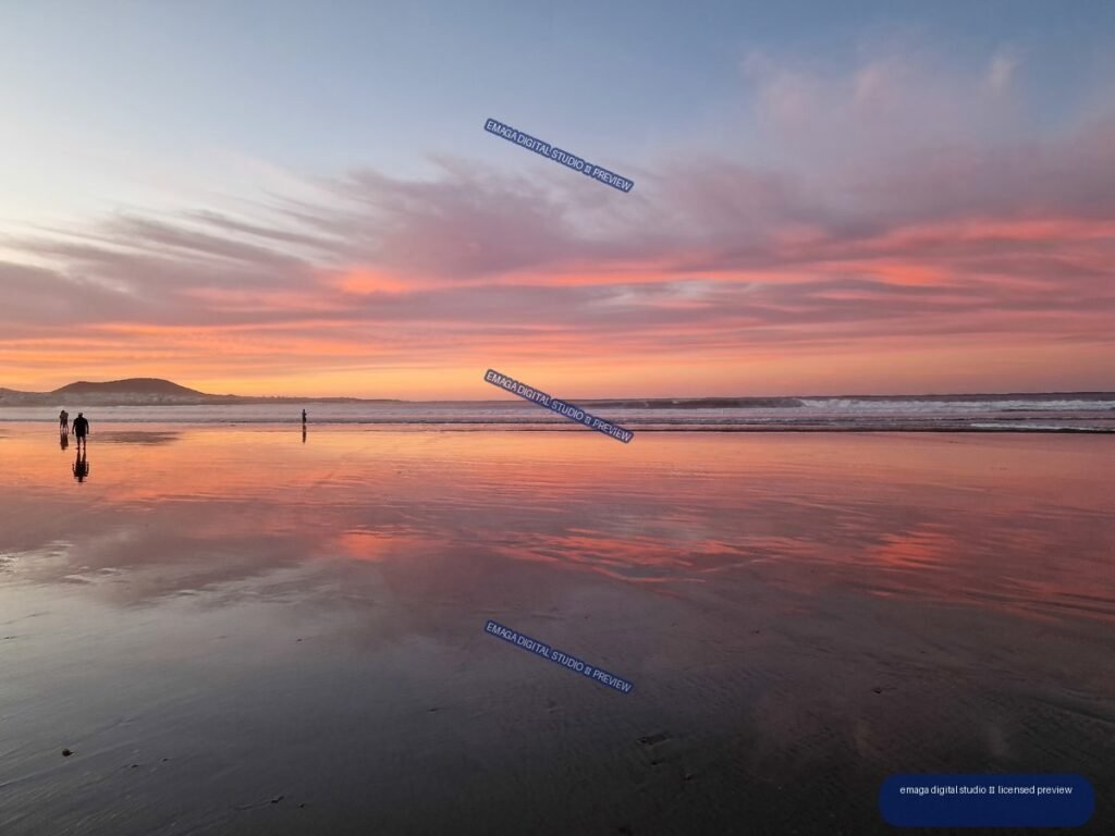 Sunset over Famara Beach with pink clouds, shallow reflective water, waves, a low headland, and three people near the shoreline.
