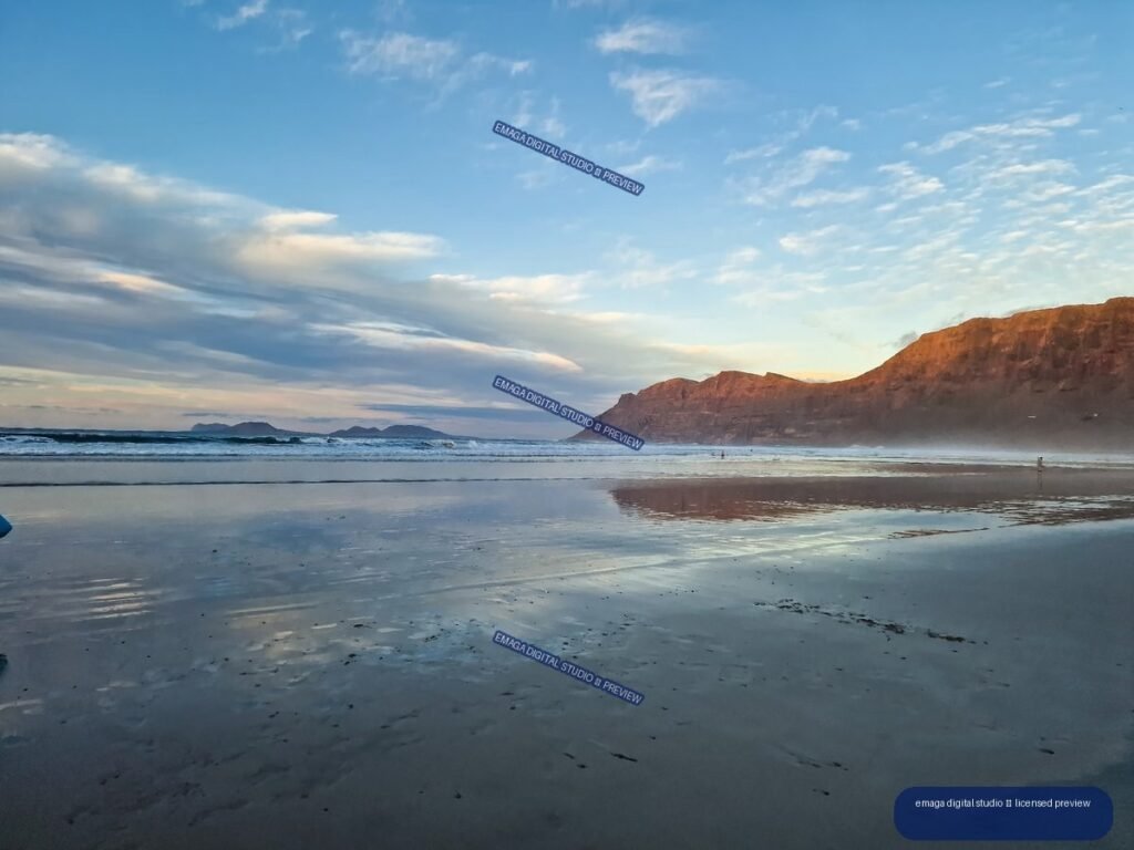 Wet reflective beach beneath reddish coastal cliffs, with gentle waves, distant rocky landforms, and a blue evening sky.