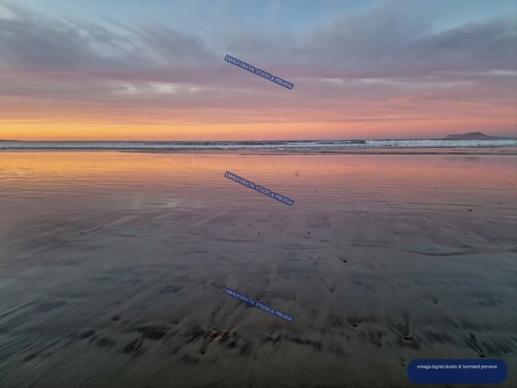Wet sand reflecting a pink, orange, and blue sunset at a beach, with gentle waves and a low landform on the horizon.