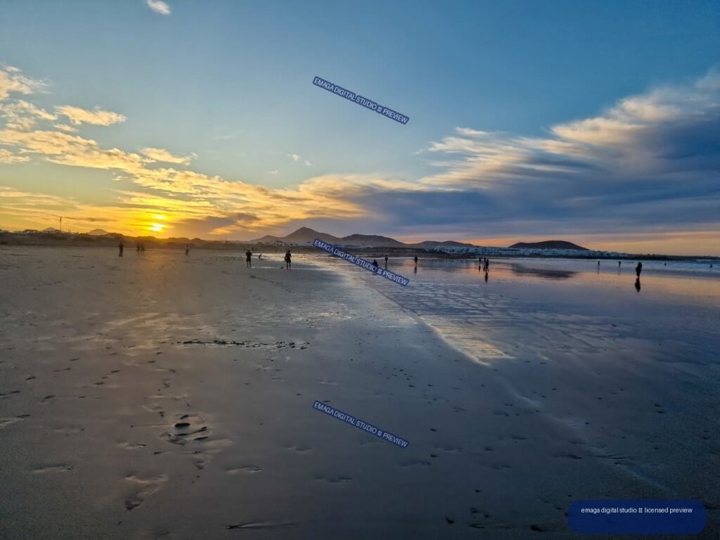 Sunset over a broad beach with volcanic hills, reflective wet sand, clouds, and distant silhouetted people.