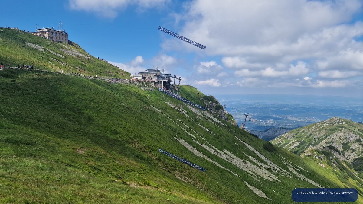 Green alpine slope with Kasprowy Wierch cableway buildings, rocky Tatra peaks, distant valley, and cloudy blue sky.