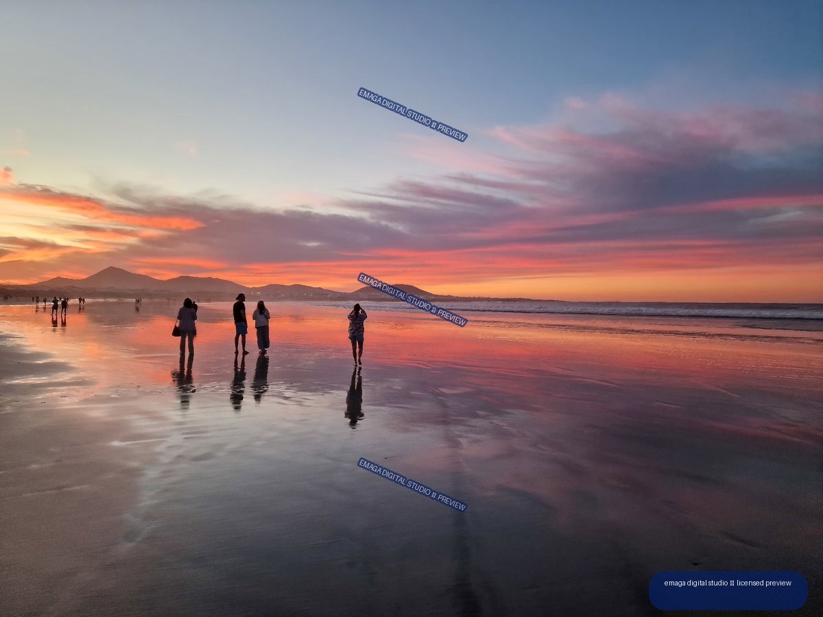 People and their reflections on wet sand at a colorful sunset, with mountains and ocean on the horizon.